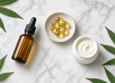 Amber dropper bottle, golden softgels in a dish and an open cream jar on marble with hemp leaves